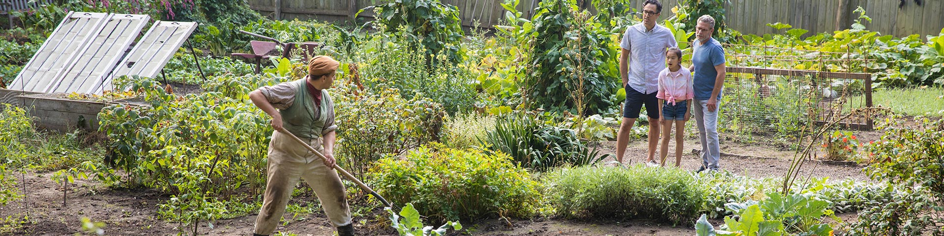 A man in period costume works in a garden as a family watches. 