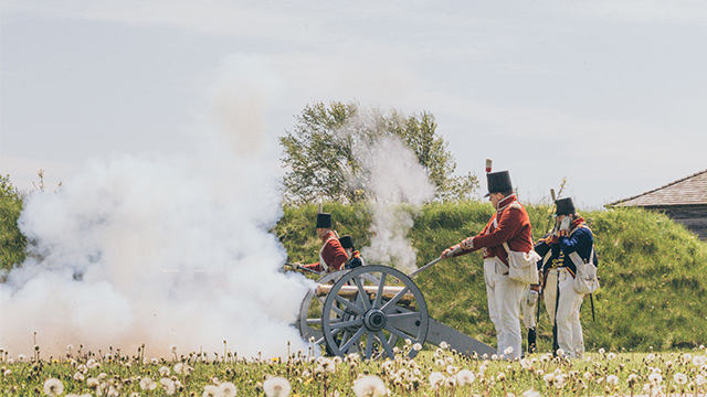 Two Parks Canada interpreters wearing historic costumes fire a cannon on a cloudy summer day.