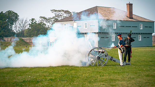 Two Parks Canada interpreters wearing historic costumes fire a cannon on a cloudy summer day. 