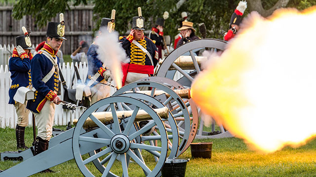 Multiple historic interpreters wearing historic costumes fire cannons during a demonstration on the grass.