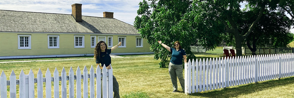 Two Parks Canada employees stand with arms outstretched in front of the officer's quarters at Fort George welcoming visitors
