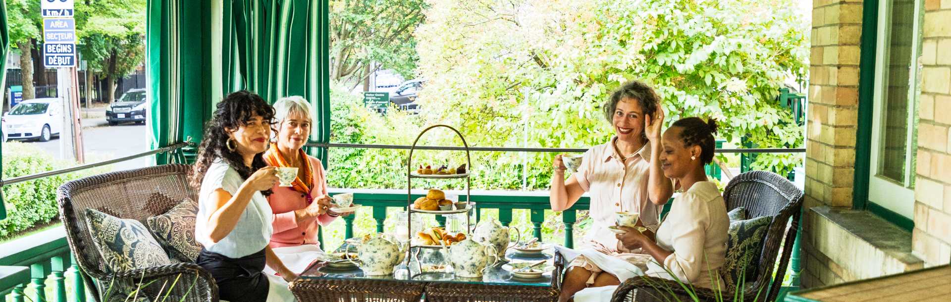 Four women enjoy tea and pastries on a green porch.