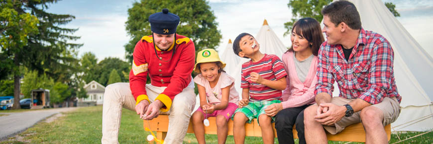 A young family with two children and two adults are sitting on a bench outside with a soldier enjoying toasting marshmallows by a fire. 