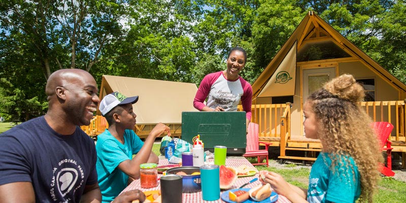 Une famille de quatre personnes sourient en prenant un repas par une journée ensoleillée à l’extérieur de leur oTENTik