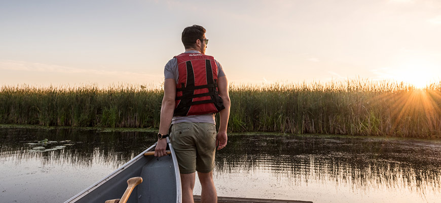 Un jeune homme transporte un canoë dans l’eau alors que le soleil se cache derrière les longues herbes du marais.