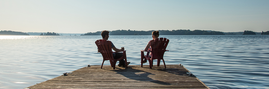 Man and woman sit on dock at edge of water in red chairs.