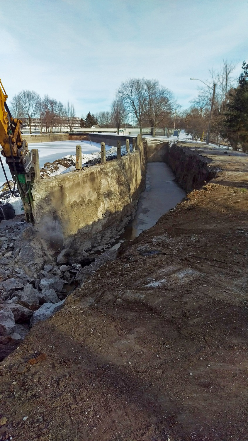 Winter. A rock breaker chips the edge of a concrete wall. The earth behind it is excavated