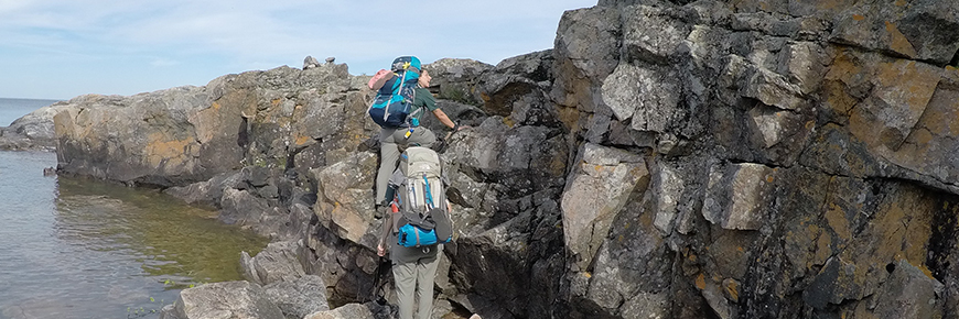 Two hikers scramble up a small rock face along Lake Superior, while carrying backpacks.