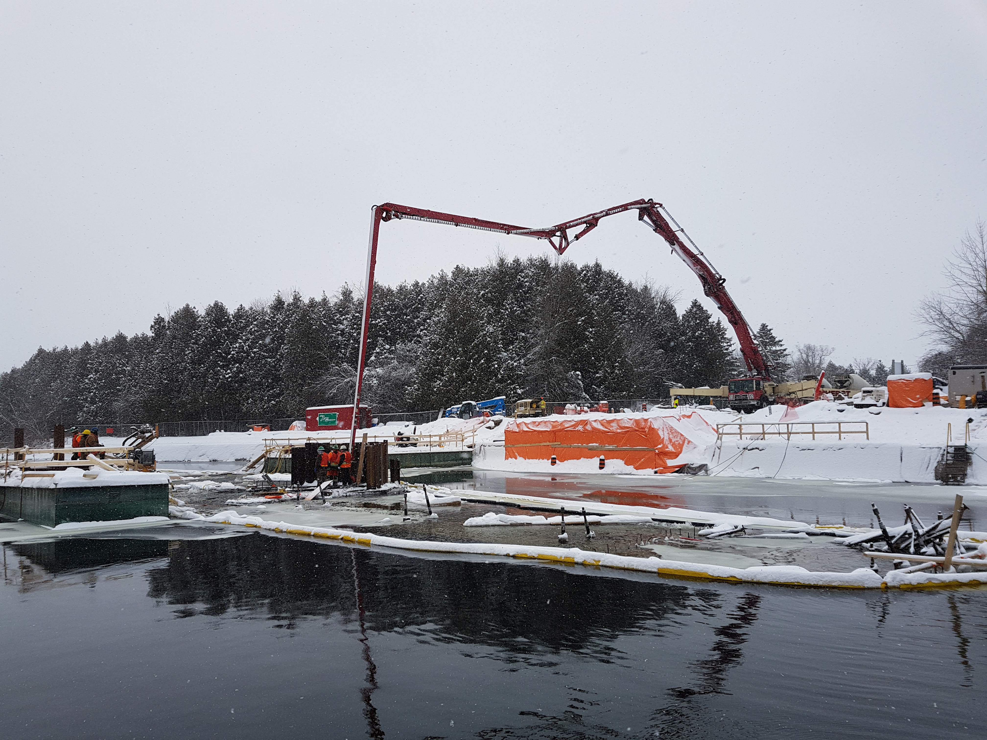 A snowy construction site. The extendable arm of a concrete pump truck reaches out over the water.