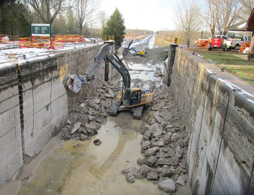 An excavator demolishes the face of a concrete wall inside a lock chamber.