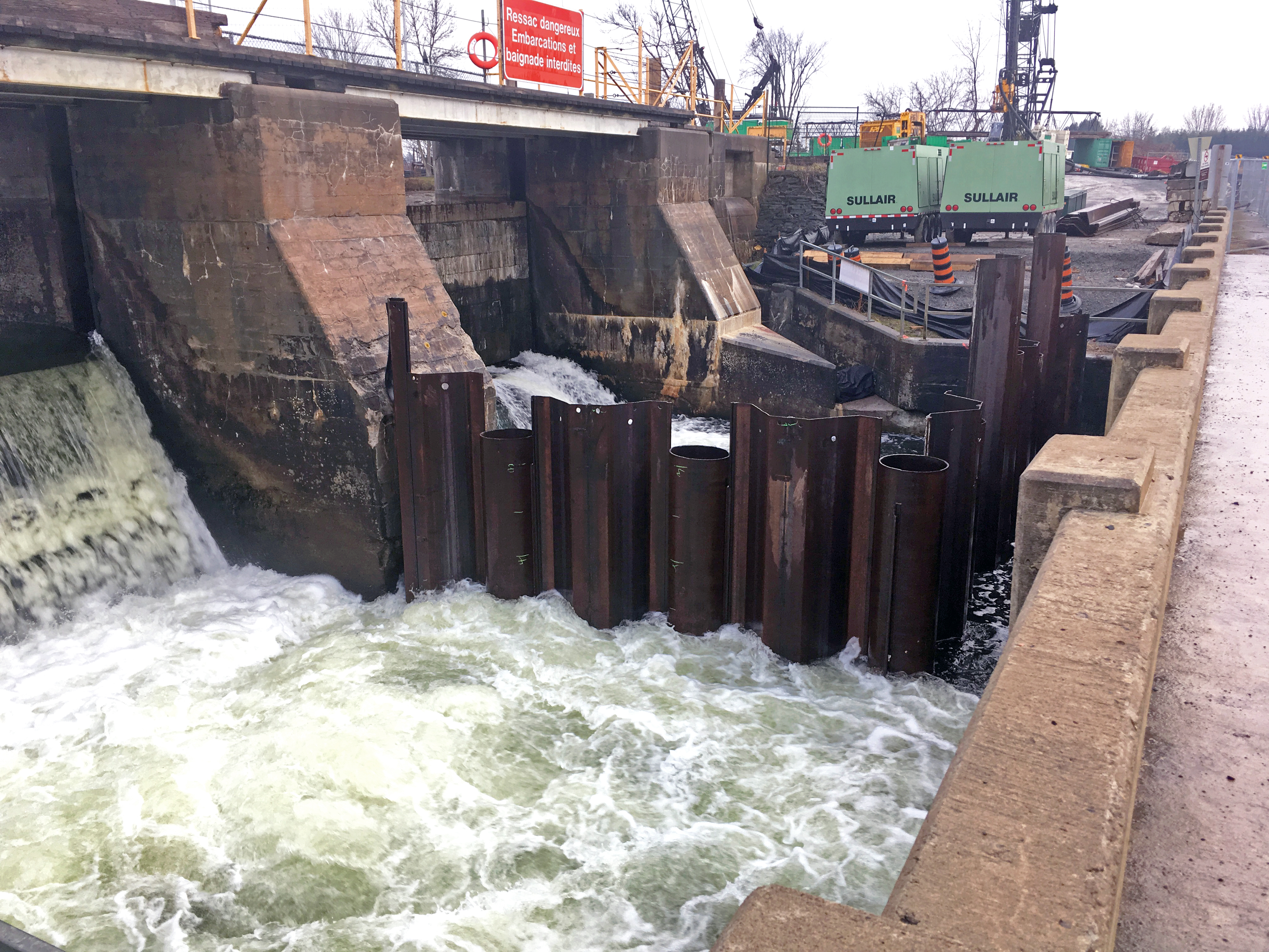 Débit sortant aux deux pertuis du barrage en béton. Des palplanches ondulées entourent le pertuis de droite et forment un carré.