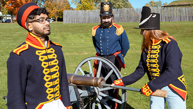 3 visitors dressed in period costume stand around an old canon as they learn how to load it.