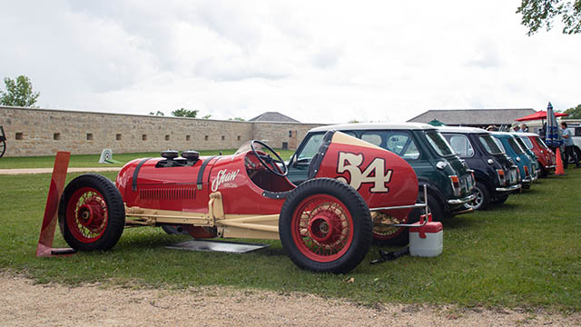 A red vintage race car with the number 54 parked on grass beside a row of classic cars at an outdoor car show