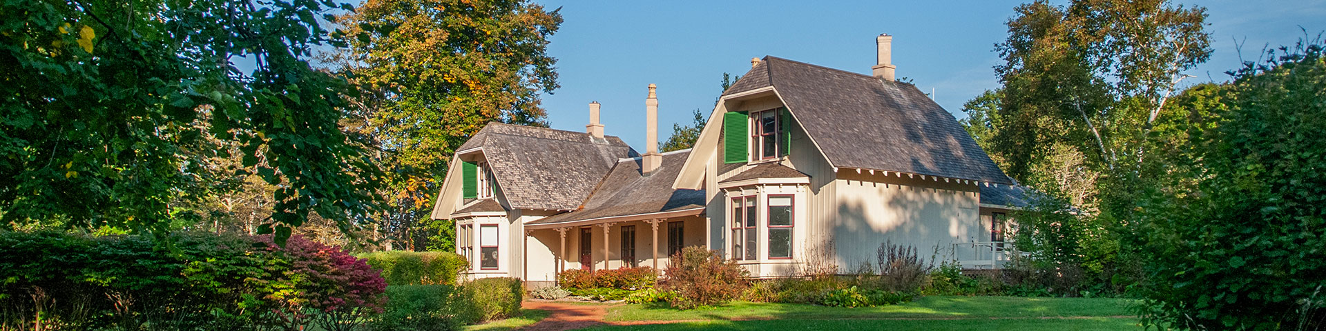 A large white older home with a black roof and many windows sits surrounded by lush green trees and gardens on a sunny day with blue skies.