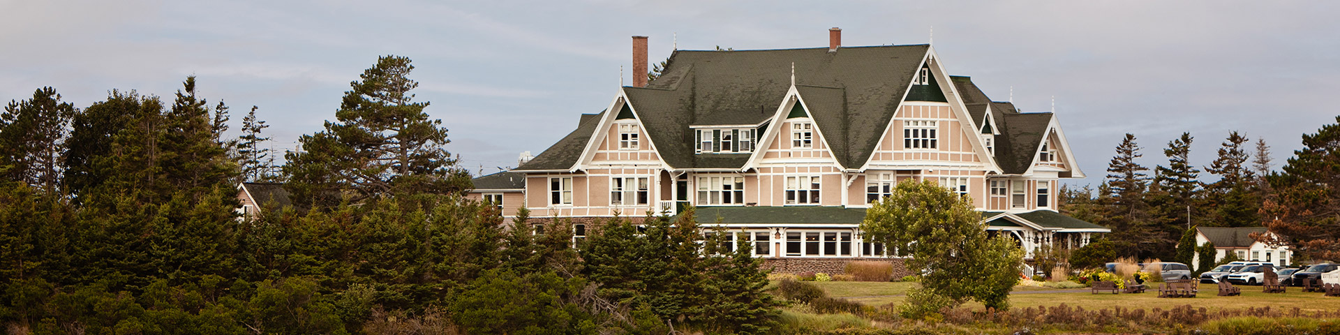 Grande maison de style victorien avec des toits à pignons raides entourée d’arbres verts denses sous un ciel nuageux.