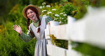 Anne waves hello from behind a white fence at Green Gables