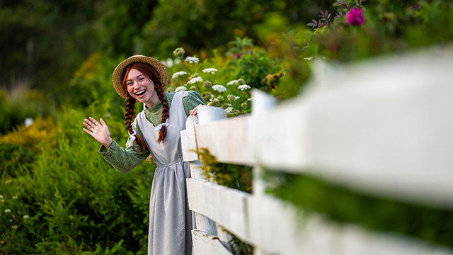 Anne nous salue de derrière une clôture au Green Gables.