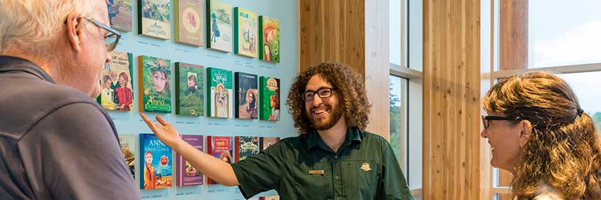 Un guide se tient devant le mur des couvertures de livres internationaux dans la salle d'exposition du centre d'accueil de Green Gables, et parle à un homme et une femme qui lui font face. 