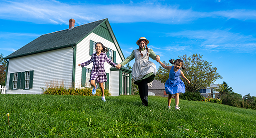 Anne runs across the lawn at Green Gables House with two young children.