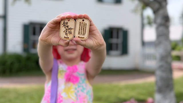 Un jeune enfant tient deux petites étiquettes en bois, debout devant une maison blanche aux moulures vertes, par une journée ensoleillée.