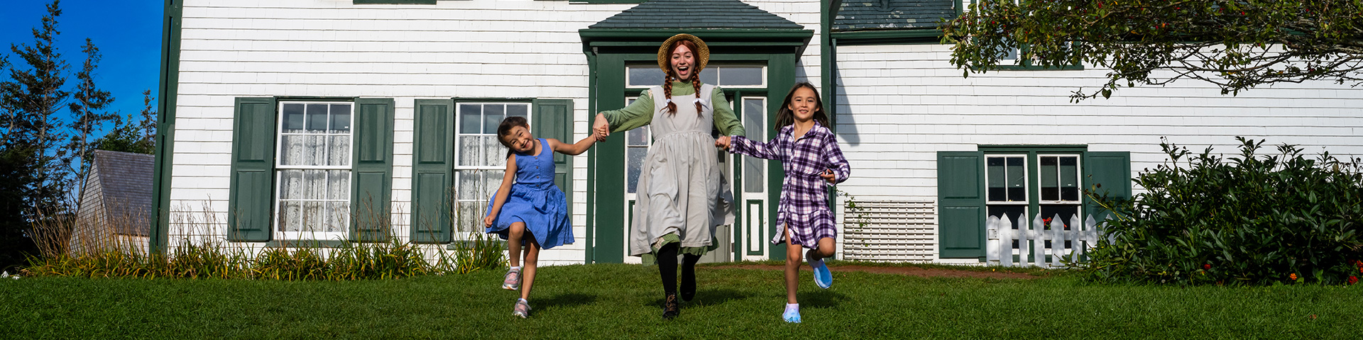 Anne runs across the front lawn of Green Gables House holding hands with two young friends.