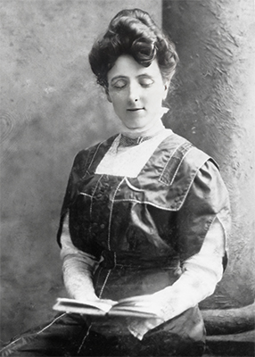 A black and white photo of LM Montgomery sitting with a book by a pillar.