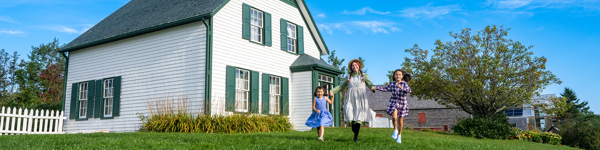 Anne and two young friends run across the lawn at Green Gables House