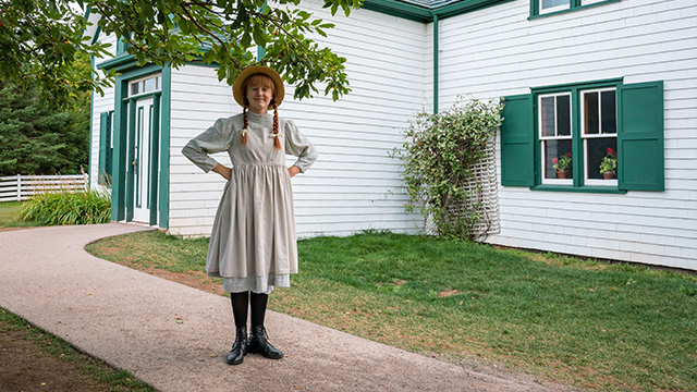 Anne stands under a tree beside a walkway that leads to Green Gables House.