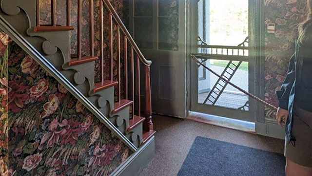 Sun shines through the open front door of Green Gables House.  There is steep stairs to the second floor. 