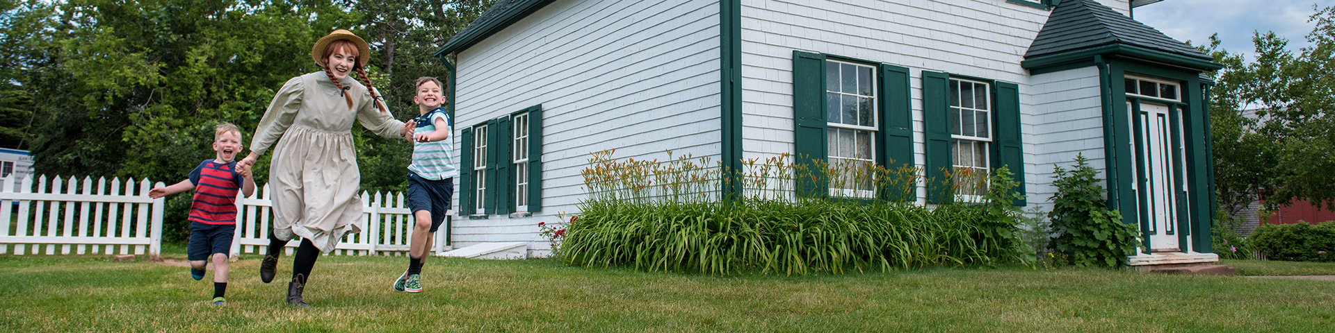 Deux jeunes garçons courent avec Anne Shirley, personnage costumé, dans les jardins du site patrimonial Green Gables, lors d'une journée d'été.