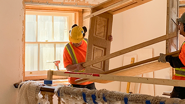 Two workers wearing a hard hat and reflective vest move a window frame into place. 