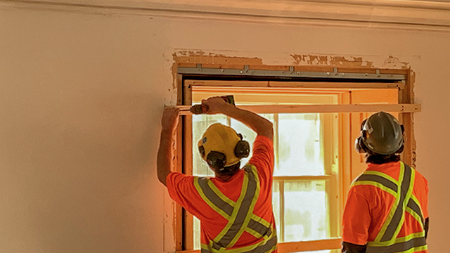 Two workers wearing hard hats and reflective vests place a window frame into place against a yellow wall.