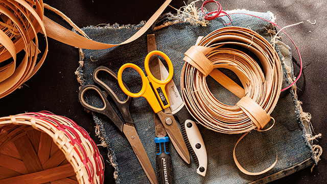 Two pairs of scissors, one yellow and one black, placed on a denim cloth alongside coiled strips of wood for basket weaving.