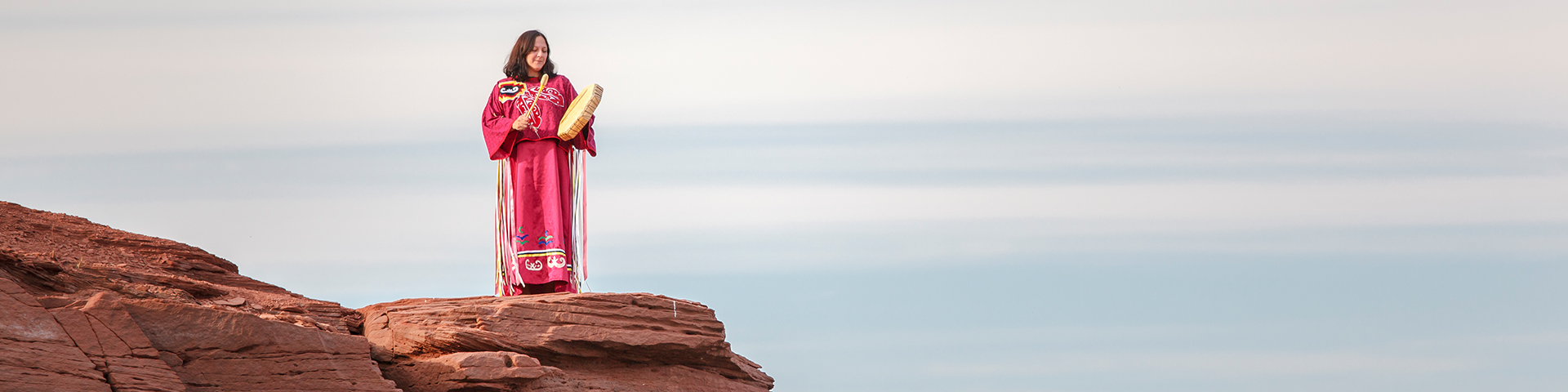 A Mi'kmaq woman plays a drum on a cliff overlooking the mouth of the Hillsborough River. Skmaqn–Port-la-Joye–Fort Amherst National Historic Site.