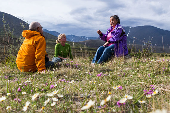 Deux personnes écoutent une femme parler dans un champ de fleurs sauvages avec des montagnes en arrière-plan au parc national Ivvavik