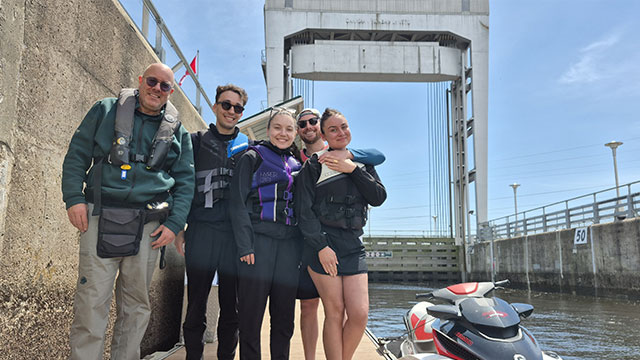 Five people wearing life jackets pose on a dock near a lock, with a jet ski beside them under a sunny