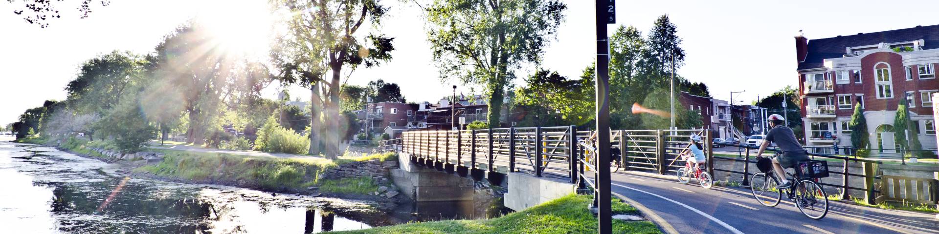 Cyclists, adult and child, on a trail along a river in an urban environment. Reflection of the sun in summer.