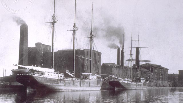 Early black and white image of a boat in a canal in front of an industrial building.