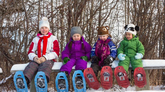 A group of children wearing snowshoes, sitting on a bench on a fine winter's day. 