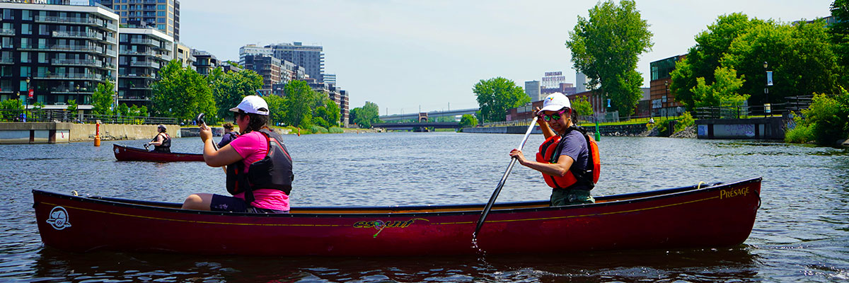 Two women in a red canoe paddling in calm water. The women are wearing life jackets and white caps. In the background, there is an urban landscape.