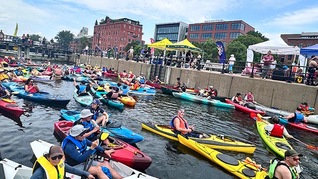 Un grand rassemblement de kayakistes navigue sur un canal animé, avec des spectateurs sur les berges.