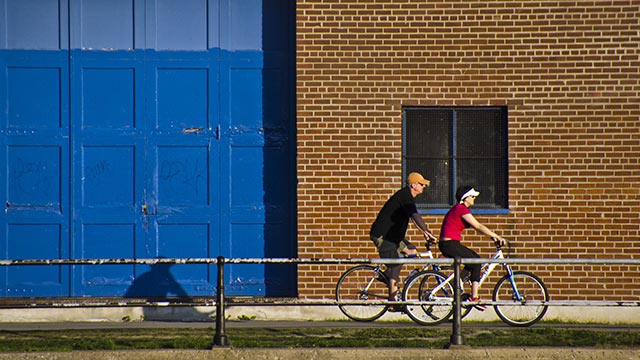 Un homme et une femme à velo, passant devant un bâtiment en brique avec une grande porte de garage bleue.