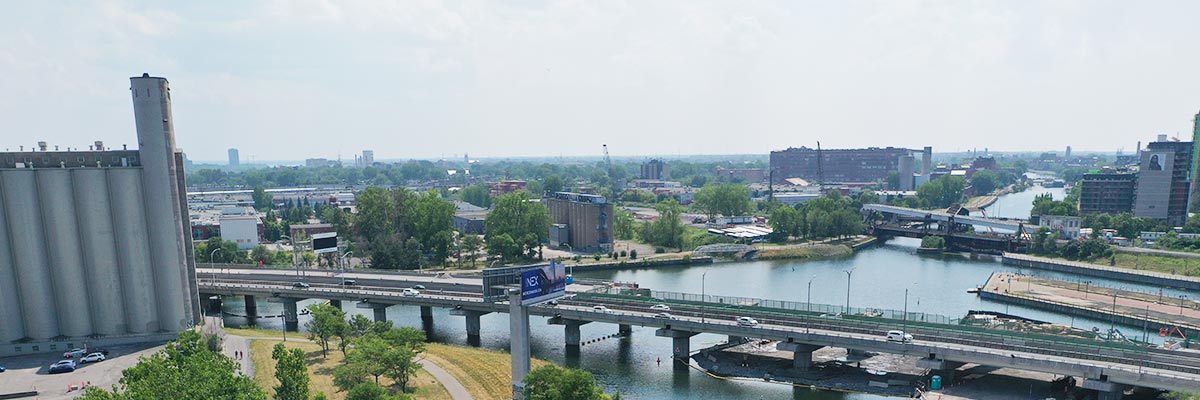 View of a canal cutting through an urban area, featuring bridges, industrial silos, and buildings surrounded by greenery.