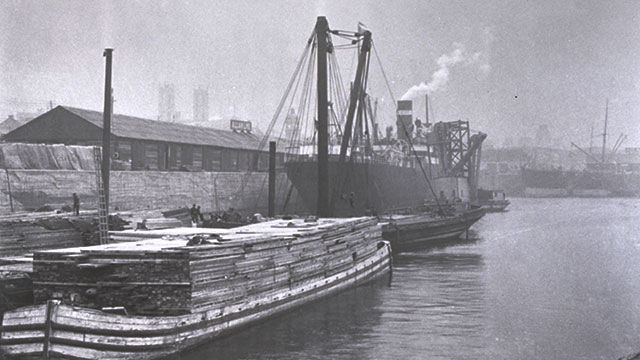 Barge chargée de bois et un navire en arrière-plan, entourés de grues et installations portuaires dans une atmosphère industrielle et brumeuse.