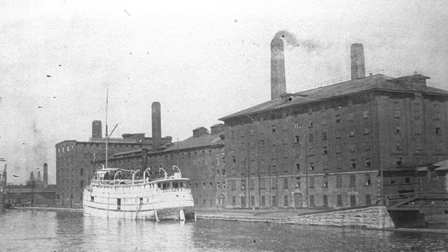 A brick industrial building by a canal, with a boat moored in front. The building, equipped with chimneys and numerous windows, evokes a historical ambiance.