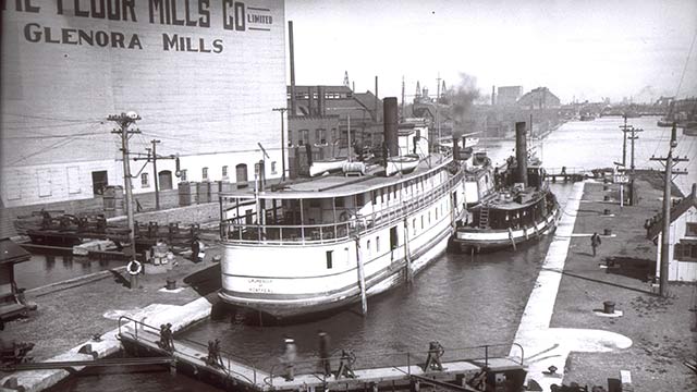 Image historique d'un quai et deux bateaux amarrés, entourés de bâtiments industriels et de personnes engagées dans des activités portuaires.