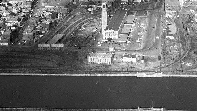 Vue aérienne en noir et blanc d'une zone industrielle avec plusieurs bâtiments, des parkings et un canal navigable au premier plan.
