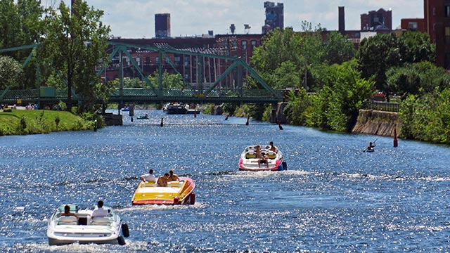 3 bateaux à moteur transportant plusieurs personnes naviguent dans un canal entouré de verdure. On aperçoit un pont routier de structure métallique et plusieurs bâtiments en arrière-plan.