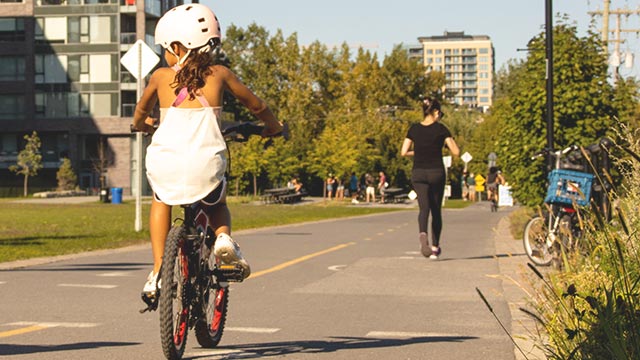 Une jeune fille portant un casque blanc fait du vélo sur une piste cyclable, avec des personnes se promenant en arrière-plan dans un espace vert.