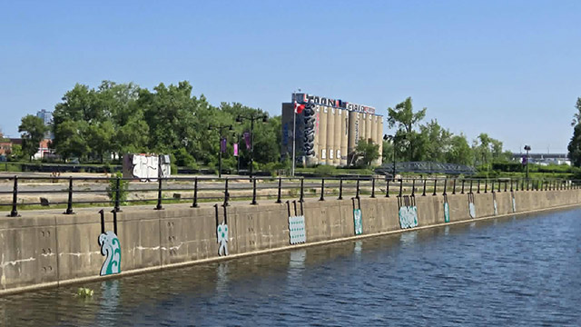 Œuvres artistiques installées sur le mur du quai du Canal-de-Lachine, avec un pont routier et des bâtiments urbains en arrière-plan.